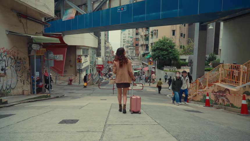 A woman with long brown hair wearing a tan jacket and a short skirt walks away from the viewer pulling a pink suitcase down a city street
