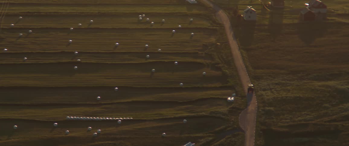 An aerial view shows a field with rows of hay bales and a dirt road running alongside it