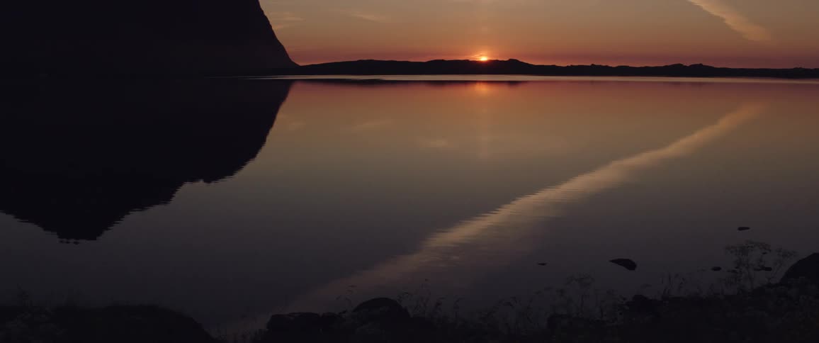 A sunset sets behind a silhouetted mountain reflecting across a calm lake in orange and purple hues with a faint horizon line and the shoreline partially visible at the edge of the frame