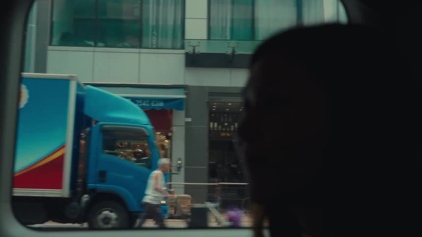 Woman riding public transportation looks out the window onto a city street lined by stores and parked delivery trucks