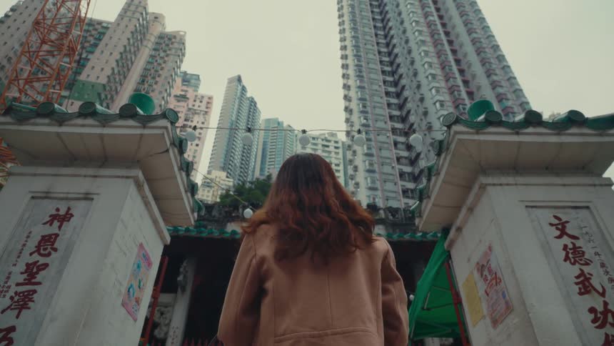 A person with long brown hair wearing a tan coat walks through the ornate gates of a temple towards a cityscape of tall buildings