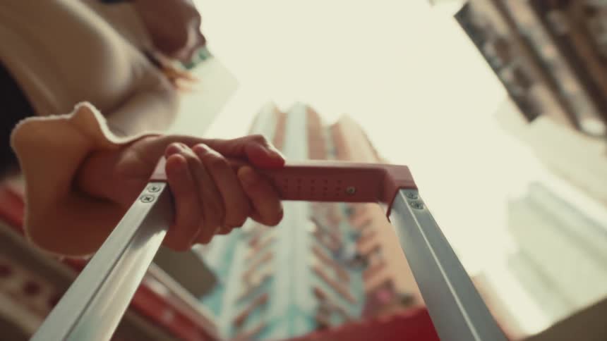 A persons hand grips the handle of a wheeled suitcase with a city scene blurred in the background