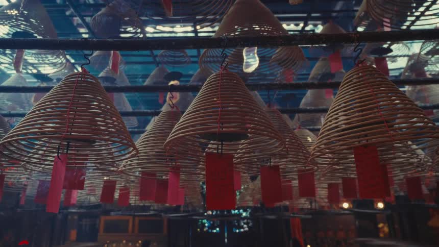 Numerous large conical spirals of slow incense hang and gracefully burn from the roof of a hong kong temple producing a soft haze with customary red pieces of paper are affixed to each coil filling the whole ceiling
