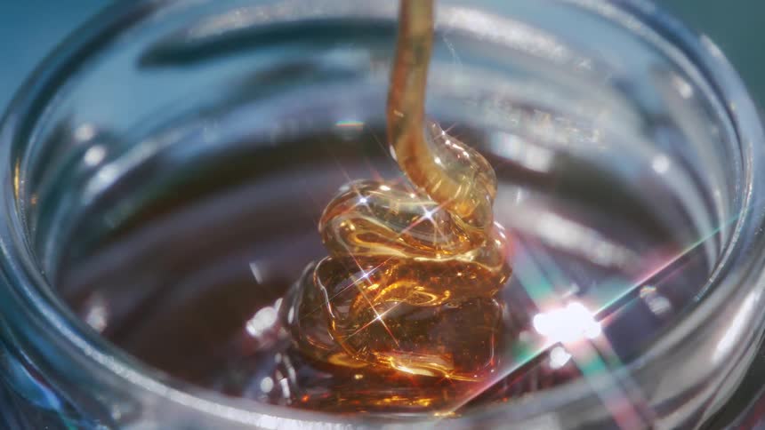 A stream of honey pours into a glass dish reflecting sunlight on a bright day