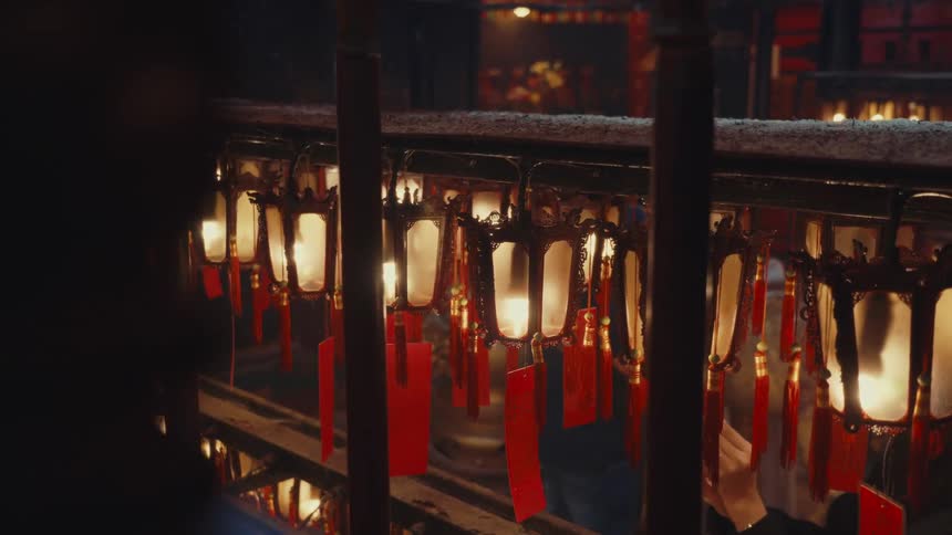 Rows of traditional chinese lanterns with glowing warm lights and inscribed wishes on red tags are on full display creating a soft ambiance within a dimly lit temple setting where people look at the laterns