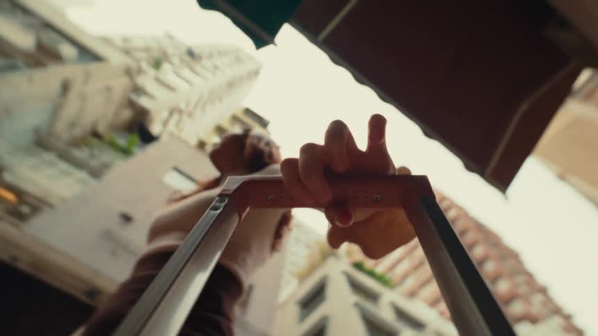 Hand holds the handle of a suitcase as the camera captures the view from above with buildings in the background
