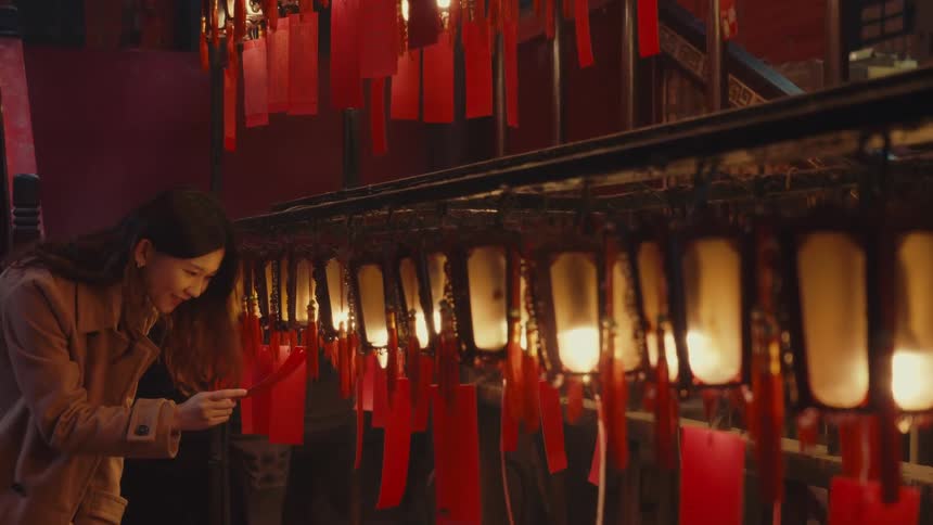 A woman in a brown coat engages with an arrangement of glowing lanterns and hanging red tags in a temple