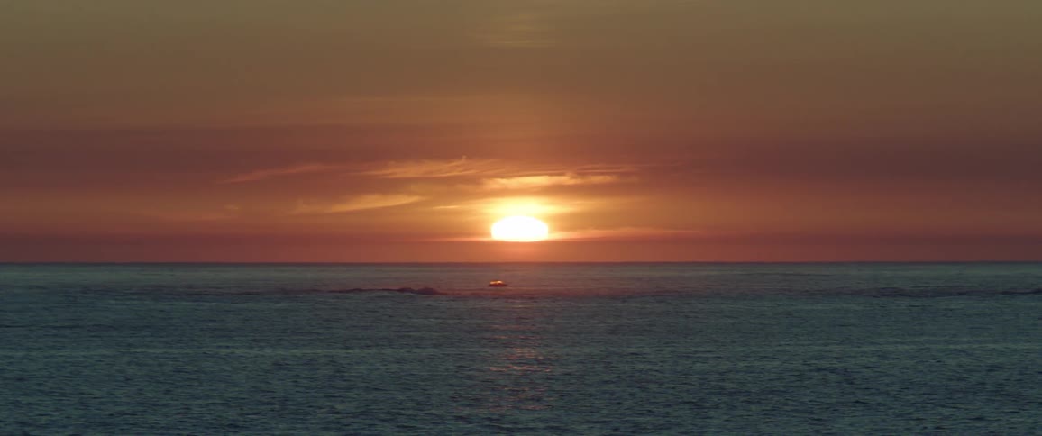 The sun sets on the horizon shining over a small boat sailing a rippled sea