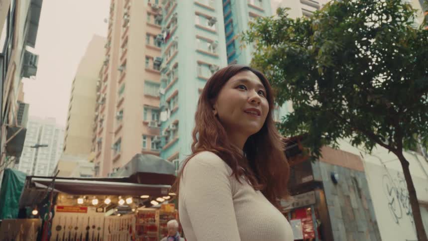 A woman with long hair stands in front of a street market with colorful items for sale surrounded by tall residential buildings and a tree