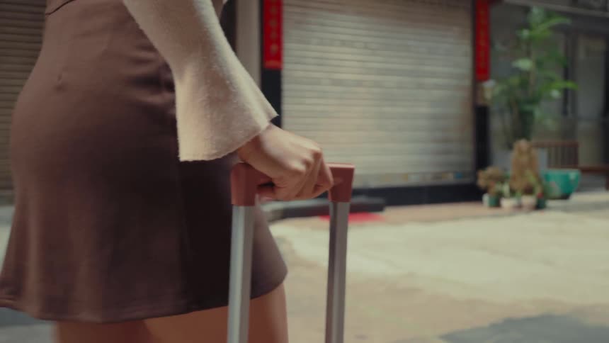 A woman in a brown skirt and white sweater pulls a silver and brown suitcase down a street with closed shops