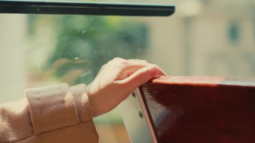 A hand in a light brown sleeve grips the edge of a reddishbrown wooden seat on a moving public transport vehicle such as a bus or tram