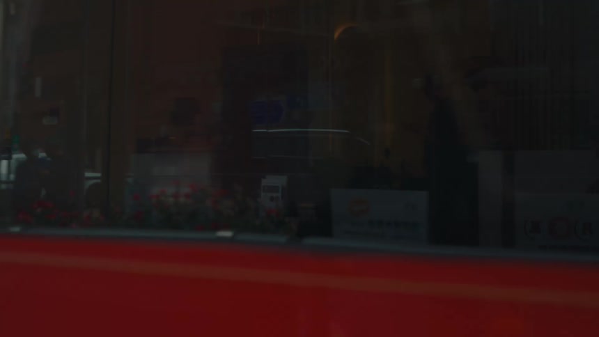 A chinese woman in a brown top and black skirt is patiently waiting at a hong kong tram station as a light blue duallevel tram approaches and a red one advances from the other direction blocking her out of view