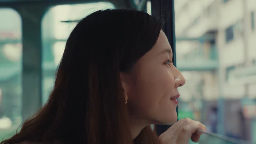 A chinese woman smiles while sitting on a hong kong doubledecker tram looking out the window as another tram passes by and she adjusts her hair