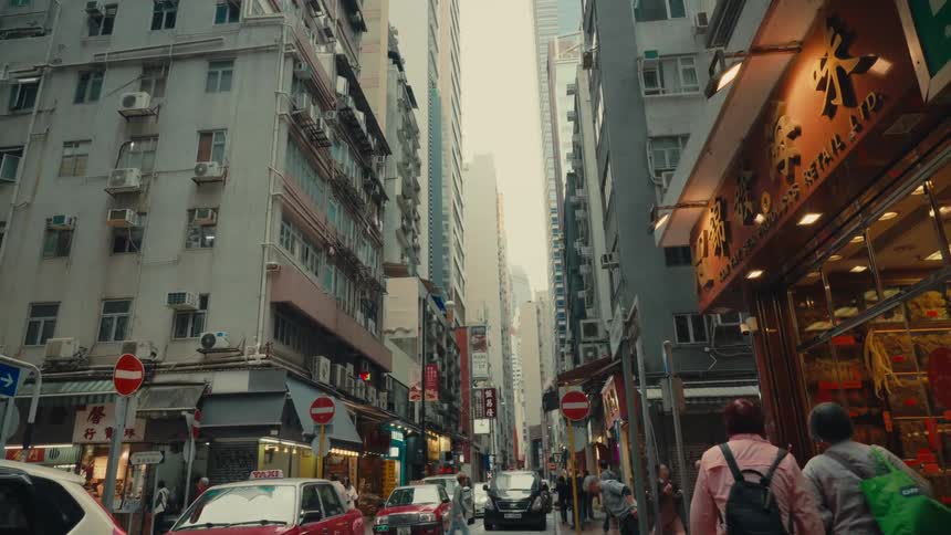 A narrow bustling hong kong street is full of foot traffic as a bunch of pedestrians navigate crowded sidewalks alongside an urban landscape lined with various shops all while the streets car traffic is stopped