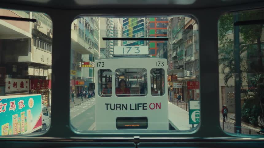 From the rear window of a hong kong doubledecker tram another tram approaches closely from behind on the same track with buildings and pedestrians visible