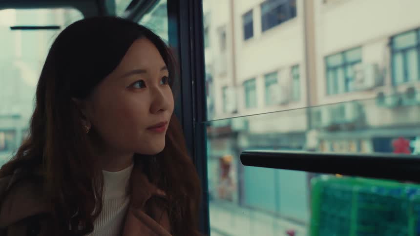 A closeup captures a woman on board a hong kong double decker tram gazing out the window beside her with an expression that slowly transforms from neutral to a smile as she takes the moving sights outside