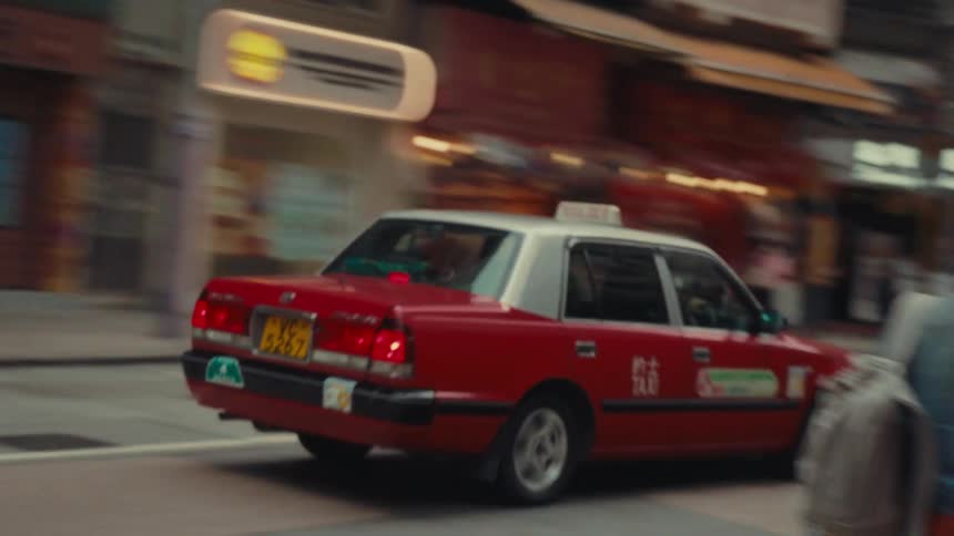An iconic red hong kong taxi makes a swift appearance moving through a busy and vibrant street scene as blurred pedestrians are seen walking on a sidewalk on the side of a busy street in hong kong