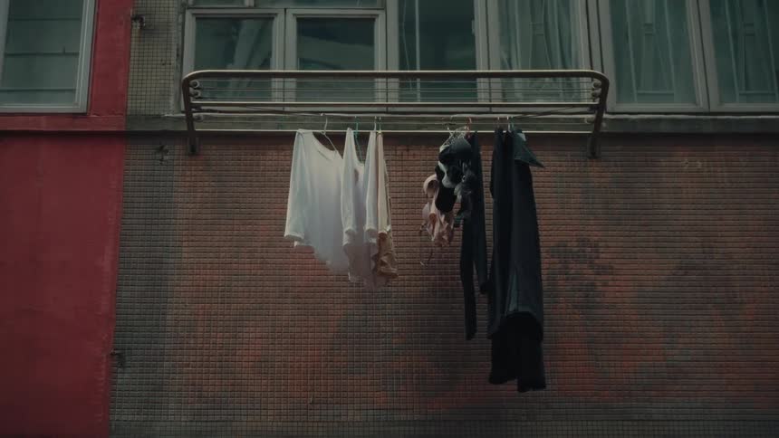Various items of clothing are hung to dry on a metal rack attached to a worn textured residential building wall in what is thought to be a narrow shadowed alleyway