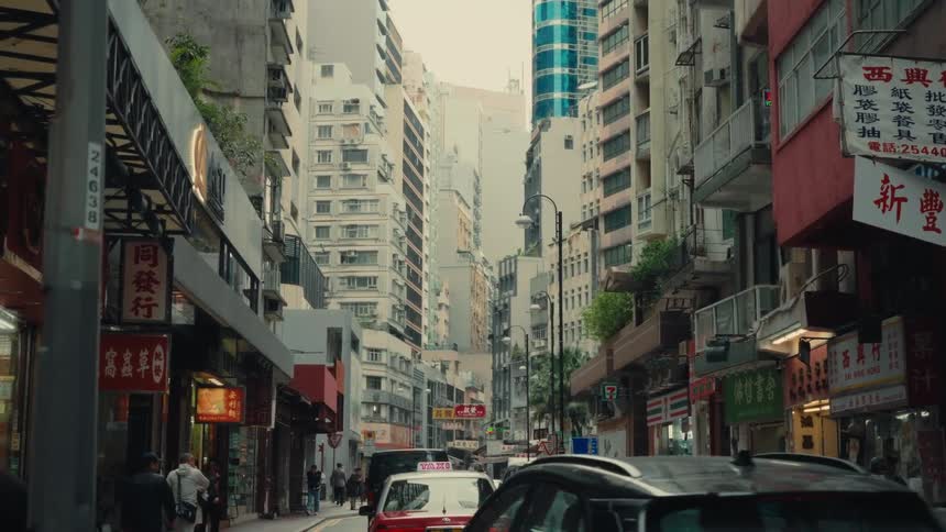A narrow city street in hong kong is depicted with densely packed buildings parked cars and pedestrians under an overcast sky