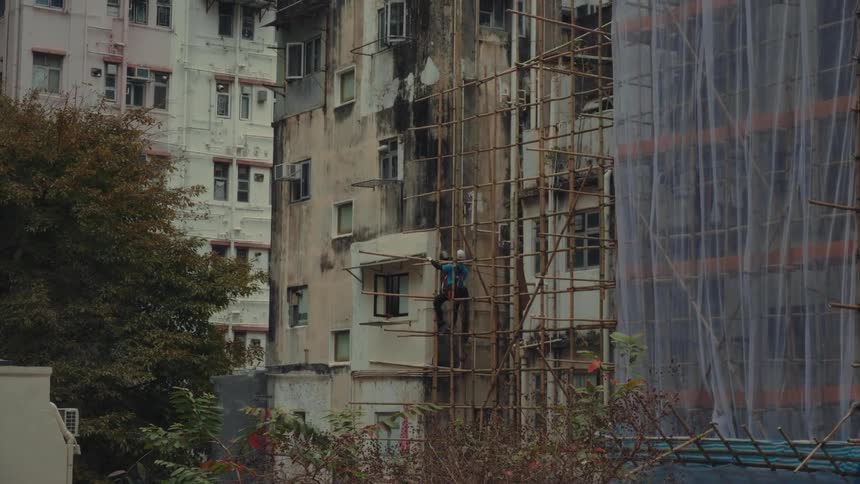 A construction worker wearing a blue shirt and white helmet climbs and works on a traditional bamboo scaffolding structure affixed to a building
