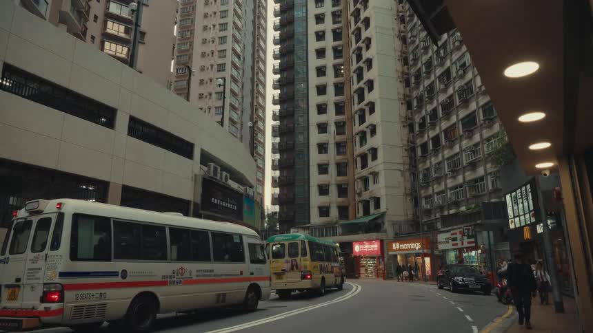 In a bustling scene a collection of hong kong people walk a busy urban road that has multiple stationary minibuses amidst towering city structures the vehicles brake signals illuminated