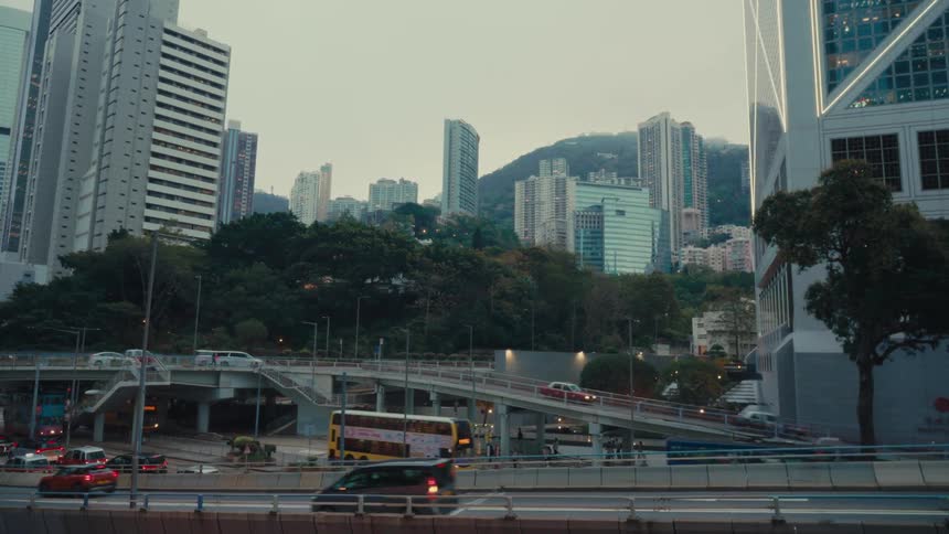 A cityscape view of hong kong shows busy streets and elevated roadways with cars and buses framed by tall buildings and greenery under a slightly overcast sky