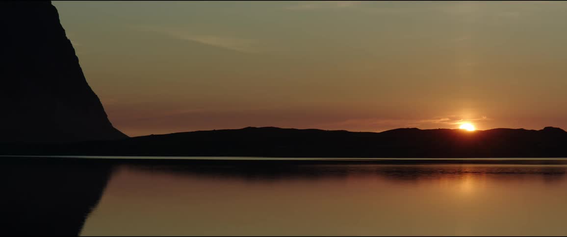 The sun dips below a distant dark ridgeline mirroring its diminishing light on the glassy surface beneath all under a soft clear sky the image gives off a nice golden glow