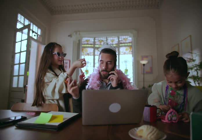 A father sits working at home with a phone and laptop while his daughters playfully dress him up and apply makeup
