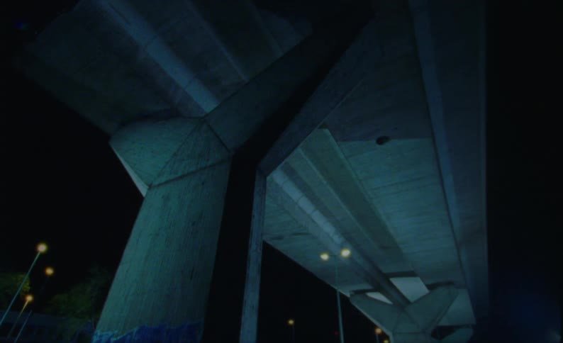 Looking upwards to reveal the underneath of a substantial concrete highway overpass the view highlights its large support structure and the expansive expanse of the road surface seen up above at night time