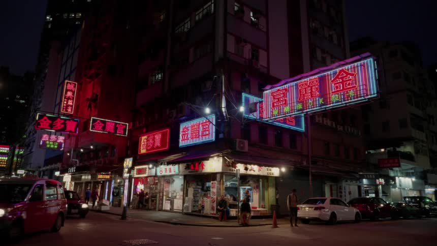 Under the glow of bright neon signs red taxis zip through a busy hong kong street corner at night building facades flicker with vibrant lights and people move along the dimly lit sidewalks while parked cars line the edges