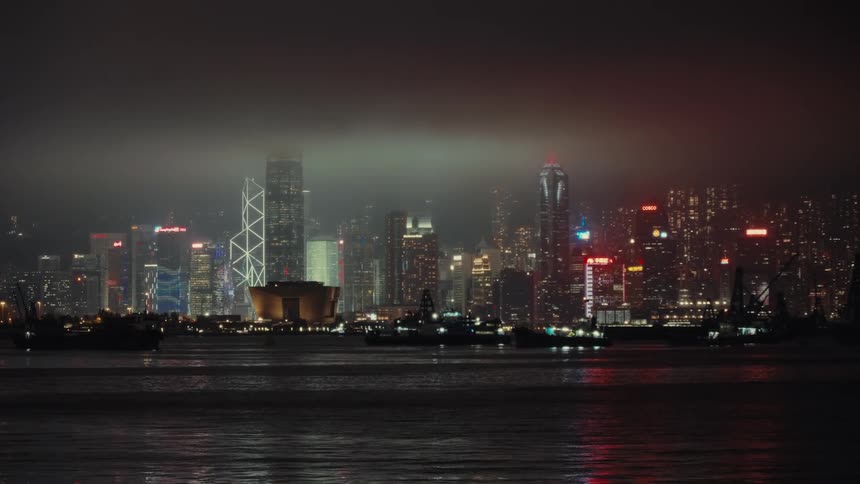 Hong Kong's dazzling metropolis illuminates the dark waters with the glow of skyscrapers while some small ships are spread throughout the bay under a dimly lit cloudy nightime sky