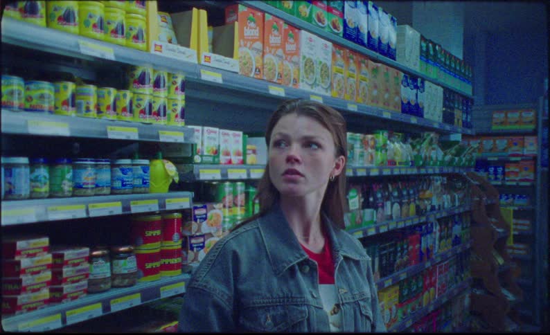 A woman wearing a denim jacket stands in a grocery store aisle stocked with canned goods boxed items and bottled beverages worriedly looks around