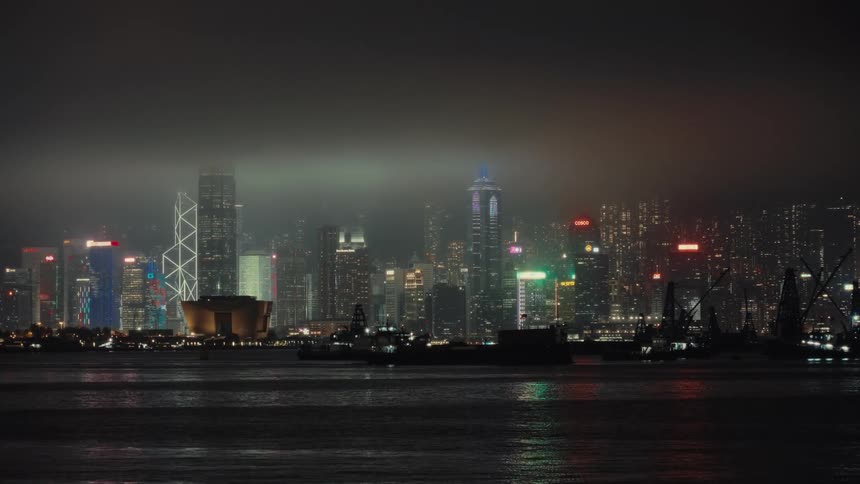 Hong Kong's dazzling metropolis skyline illuminates the waters of the bay with the glow of skyscrapers being silhouetted as a dark barge glides by under a hazy sky