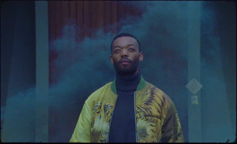 A man in a yellow jacket adorned with tiger patterns walks surrounded by swirling black smoke