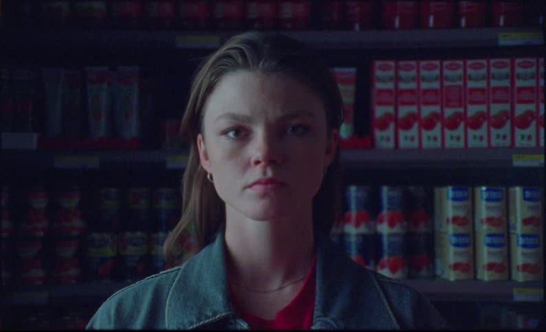 Woman standing near shelves of canned goods stares into camera while brightening red lighting shines on her face