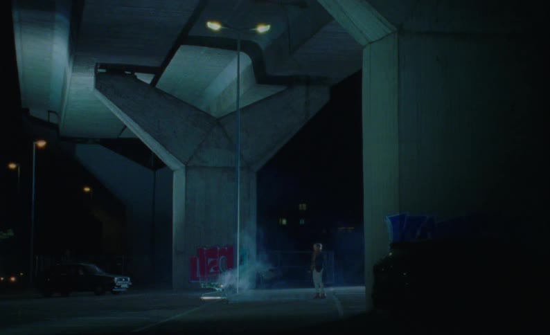 Under a concrete bridge at night a woman pushes a shopping cart with a plume of smoke originating from the cart a black car passes by