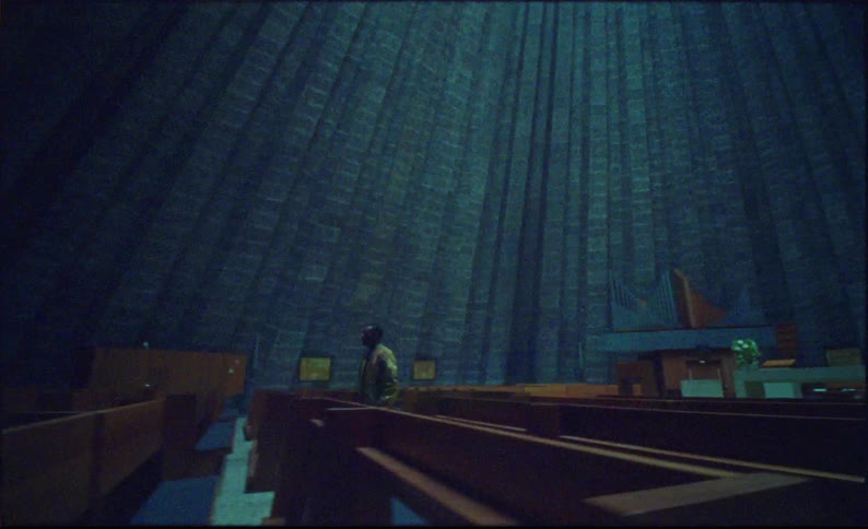 Man standing in a cathedral with vertical slanting panels turns to walk away from the altar and a tall cross