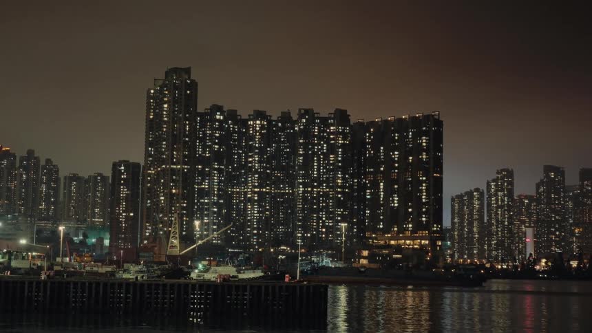 A nighttime cityscape shows densely packed high rise buildings illuminated with numerous lights reflected in the dark water of a harbor with industrial structures along the waterfront