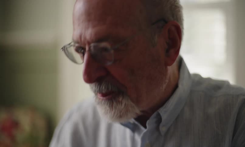A closeup captures the face of a mature man with glasses and a beard in a sunny room