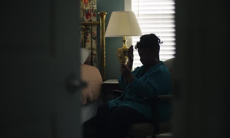 A woman sits in a dimly lit cozy room applying makeup at a mirror near a window as seen from a doorway