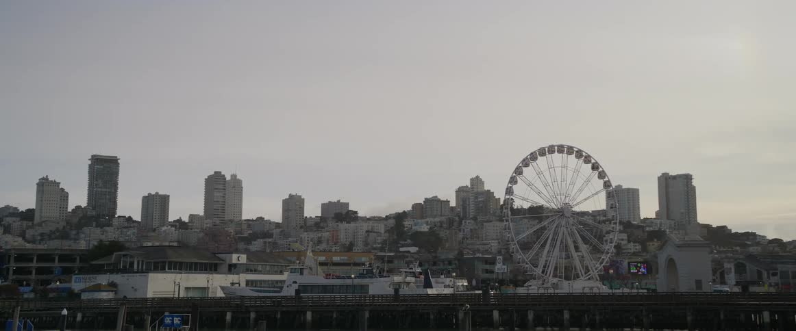 A large white ferris wheel rotates on san franciscos lively waterfront with the backdrop including a row of buildings and a vast partly cloudy sky