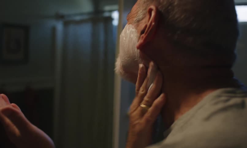 A mature man applies shaving soap to his face in front of a bathroom mirror preparing to shave his beard
