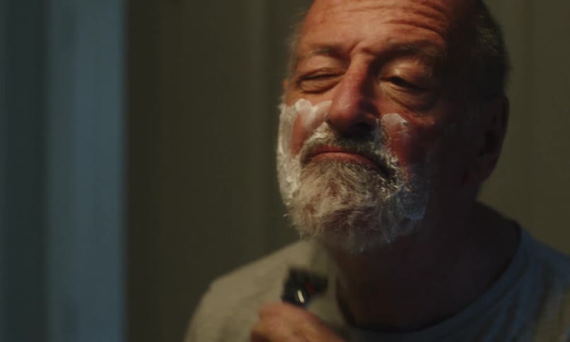 An adult man shaves his beard while looking into a mirror in a bathroom at home