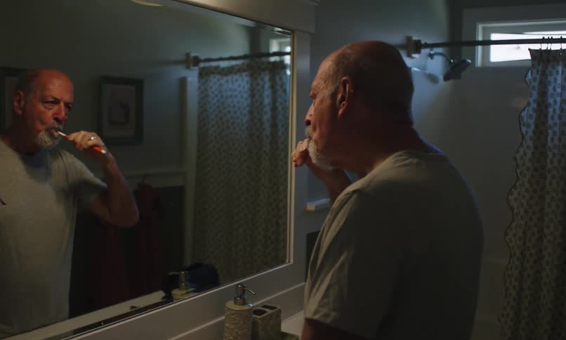 A man with a shaved head brushes his teeth in front of a bathroom mirror
