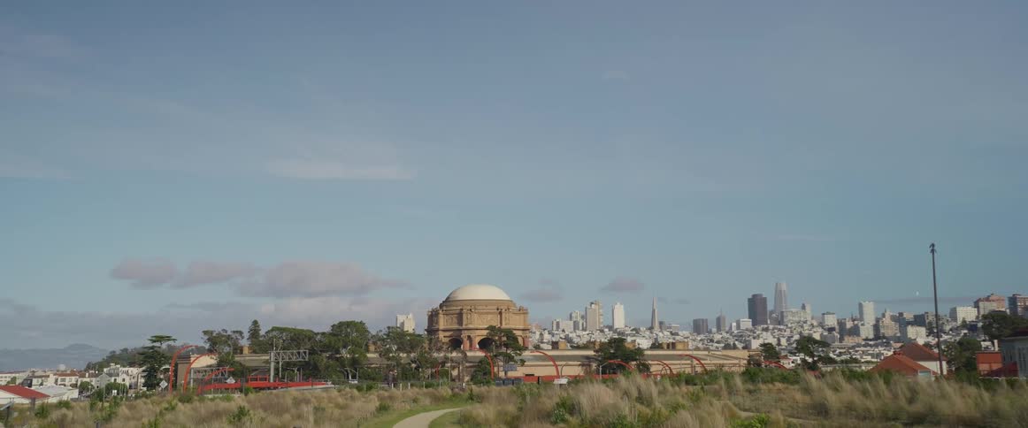 San franciscos cityscape unfolds from an elevated vantage highlighting the iconic dome of the palace of fine arts in the center with small cars traversing its nearby small paths with the larger city in the back
