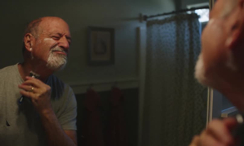 A mature man shaves his beard in front of a bathroom mirror at home