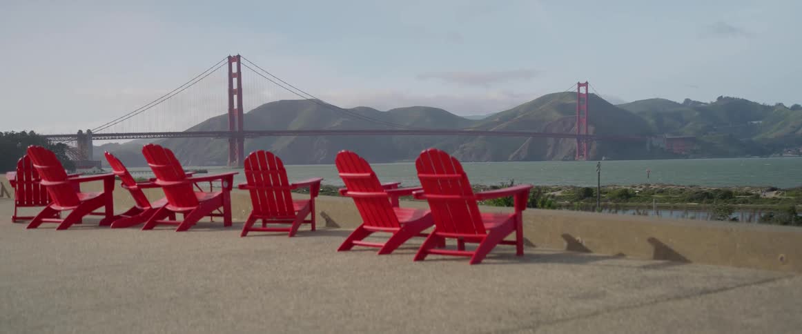 A series of bright empty red adirondack chairs are lined up along the shoreline creating a vivid contrast with the iconic golden gate bridge in the background stretching across the san francisco waterfront