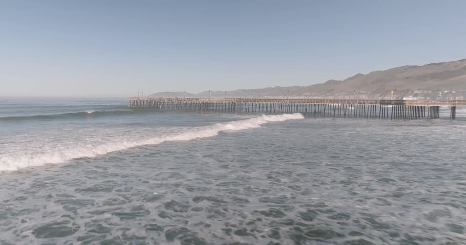 An aerial perspective moves across the sandy shoreline towards the expansive ocean it showcases the steady arrival of waves breaking into a foamy display as they meet the golden coast under a cloudy sky