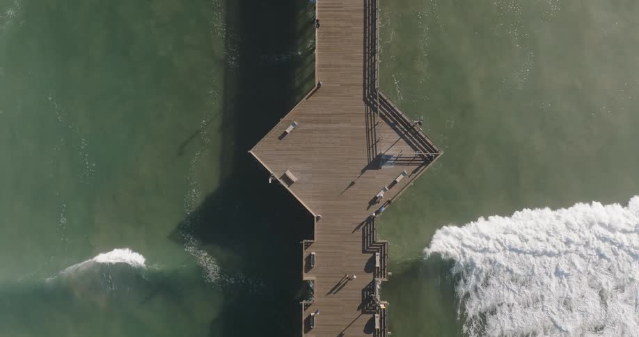 An aerial view shows a wooden pier extending into the ocean with waves crashing on either side