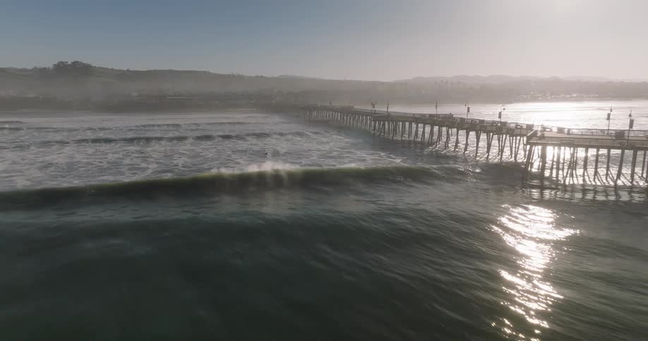 An aerial view shows a long pier extending into the ocean with waves crashing against its sides and a town visible in the background